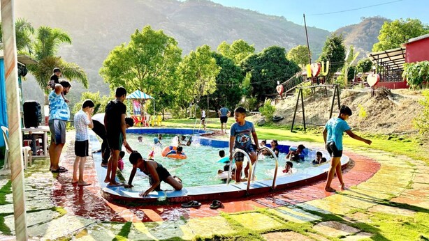 Una piscina con gente alrededor, rodeada de vegetación y colinas al fondo.