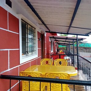 Un espacio al aire libre techado con una mesa y sillas amarillas, un edificio rojo con detalles blancos y una barandilla de metal.