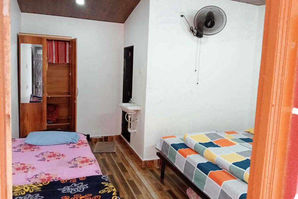 Une petite pièce chaleureuse avec un plafond en bois, un lit simple recouvert d’une courtepointe colorée, une table de chevet, un petit lavabo blanc et un ventilateur fixé au mur.