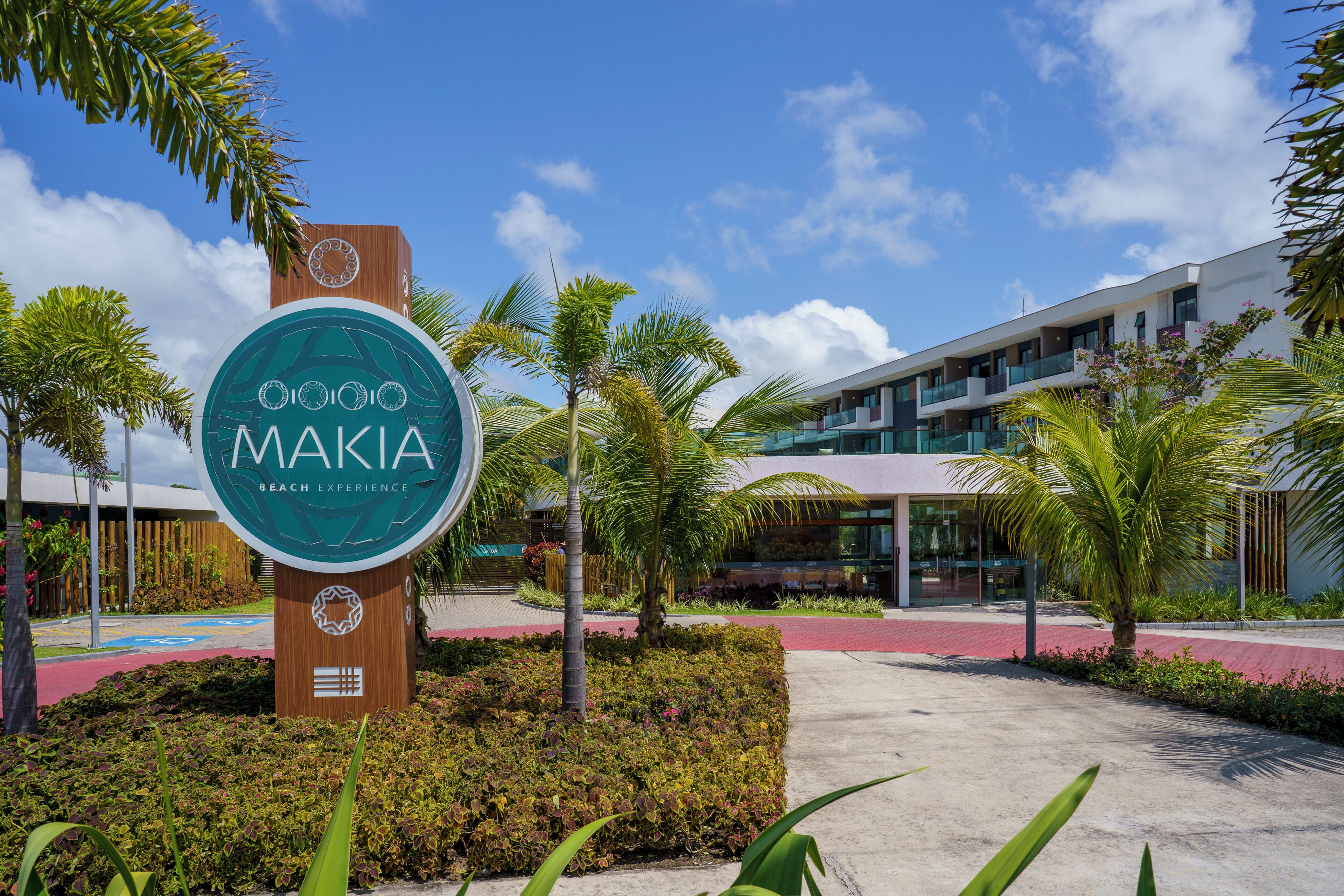A modern building with a large sign reading 'MAKIA BEACH EXPERIENCE' surrounded by greenery and palm trees.
