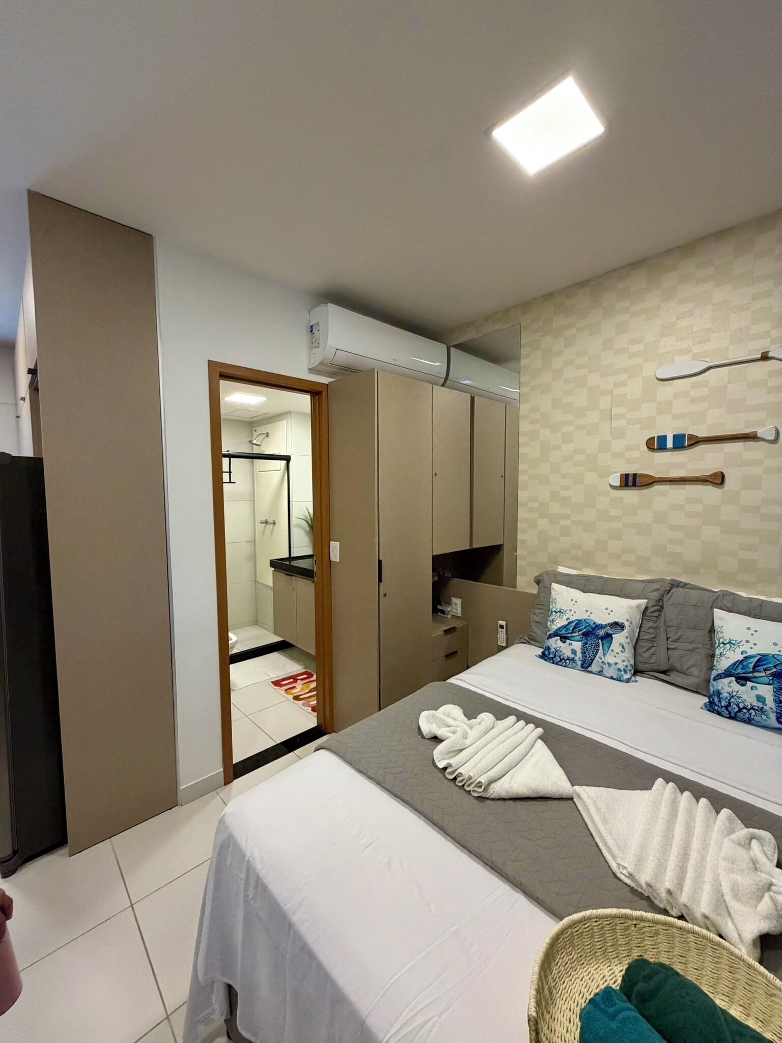 A hotel room with a bed, a wardrobe, a bathroom visible through an open door, and a ceiling-mounted air conditioner.