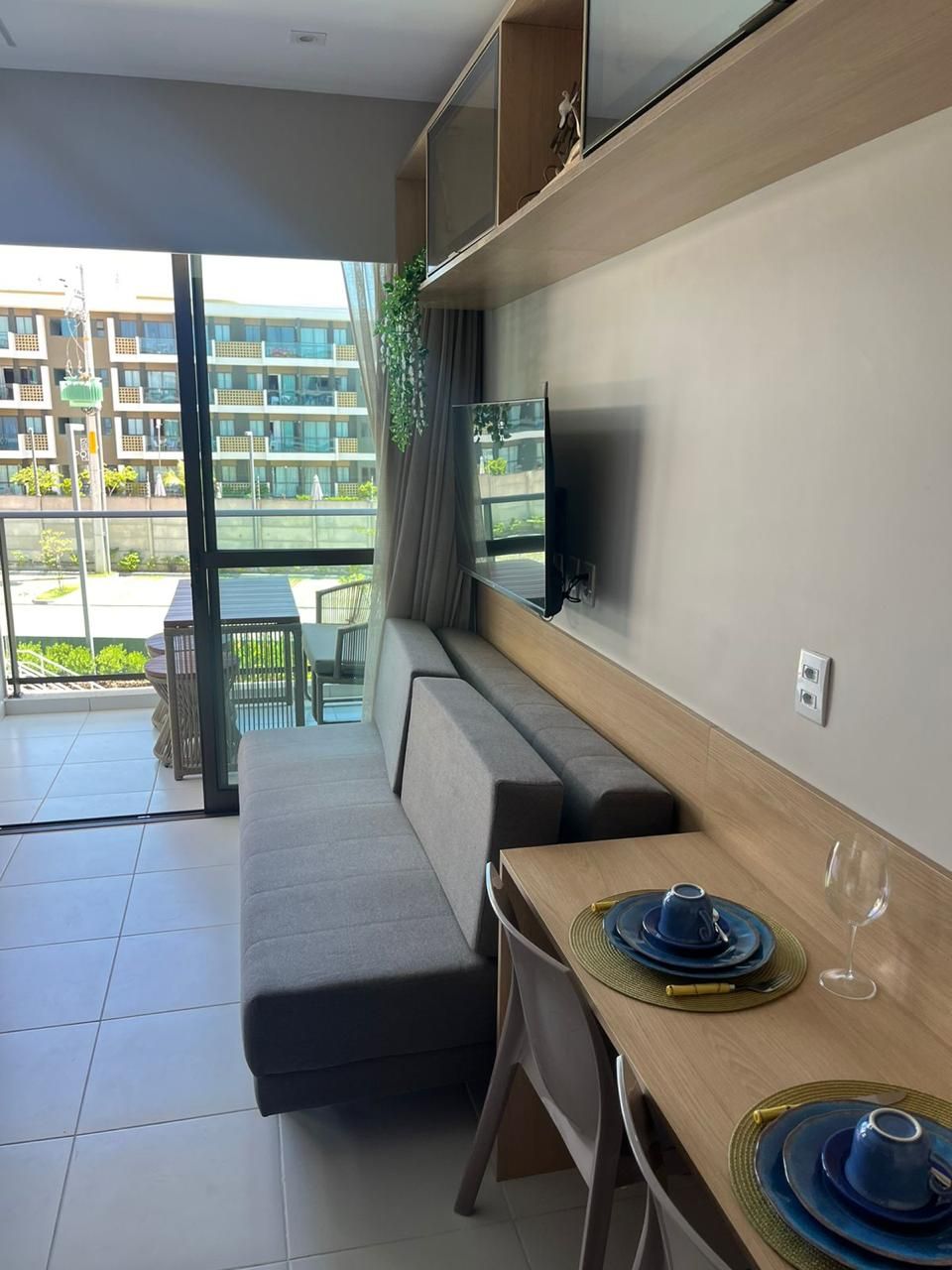 A modern living room with a sofa, a dining table set with blue plates and a wine glass, and a TV mounted on the wall.