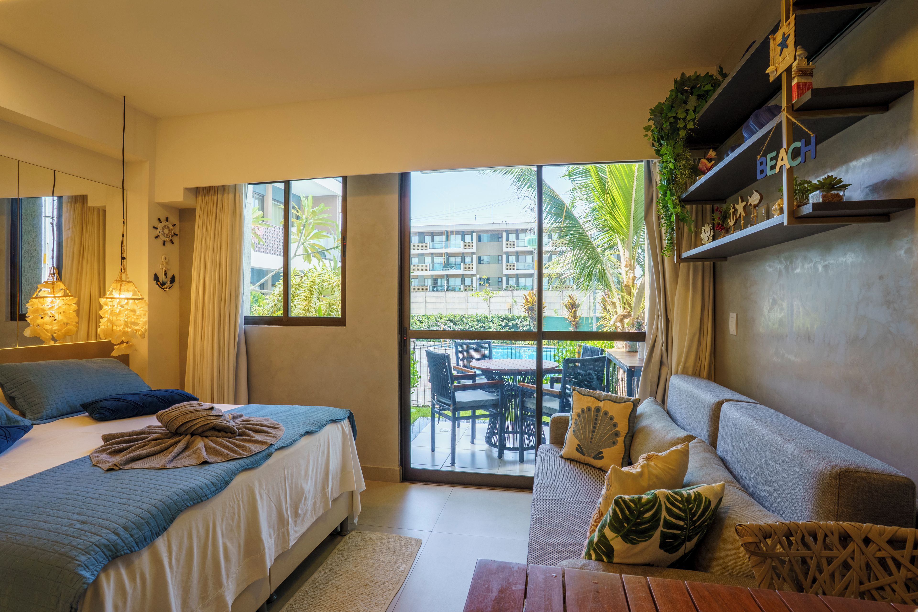 A hotel room with a bed, a sofa, a dining table, and a view of a building and greenery outside.