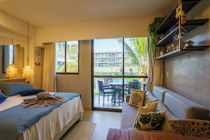 A hotel room with a bed, a sofa, a dining table, and a view of a building and greenery outside.