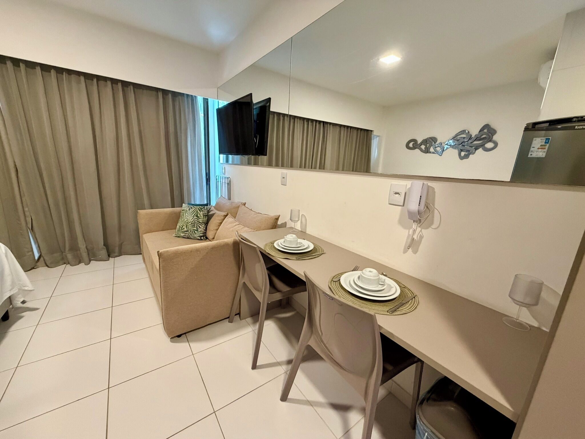 A modern hotel room with a dining area, a sofa, a flat-screen TV, a telephone, and a refrigerator.