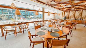 En rymlig restaurang med träbord och stolar, hängande lampor och stora fönster som erbjuder utsikt över omgivningen.