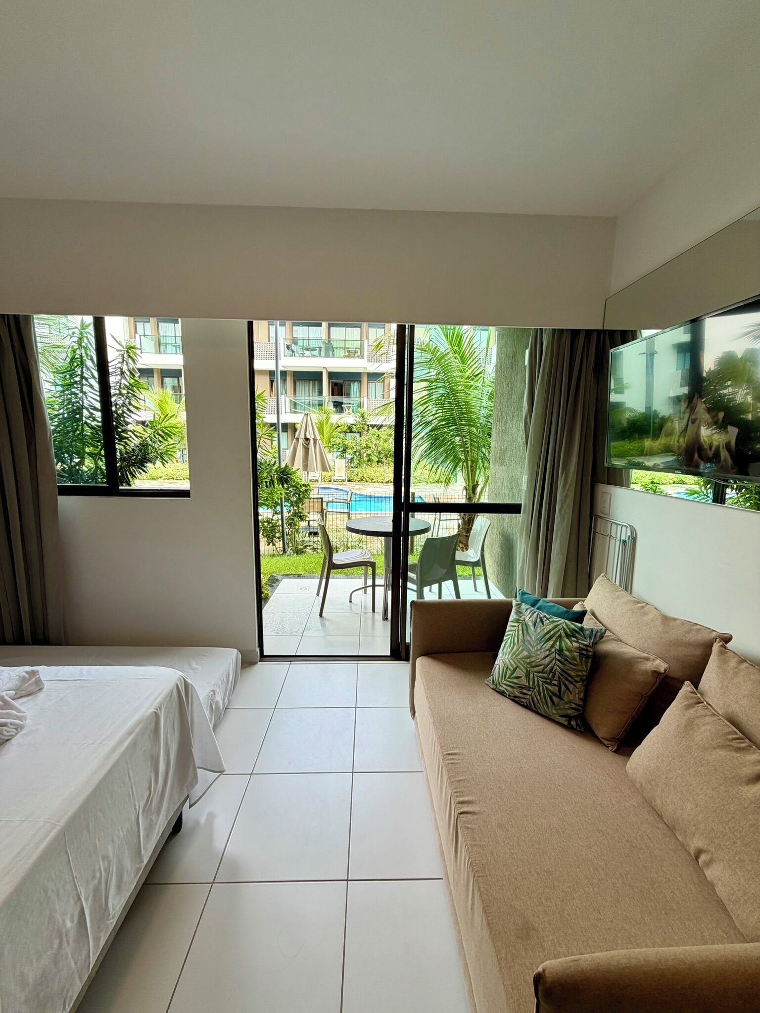 Habitación de hotel con cama, sofá, televisión y vistas a una piscina exterior y zonas verdes.