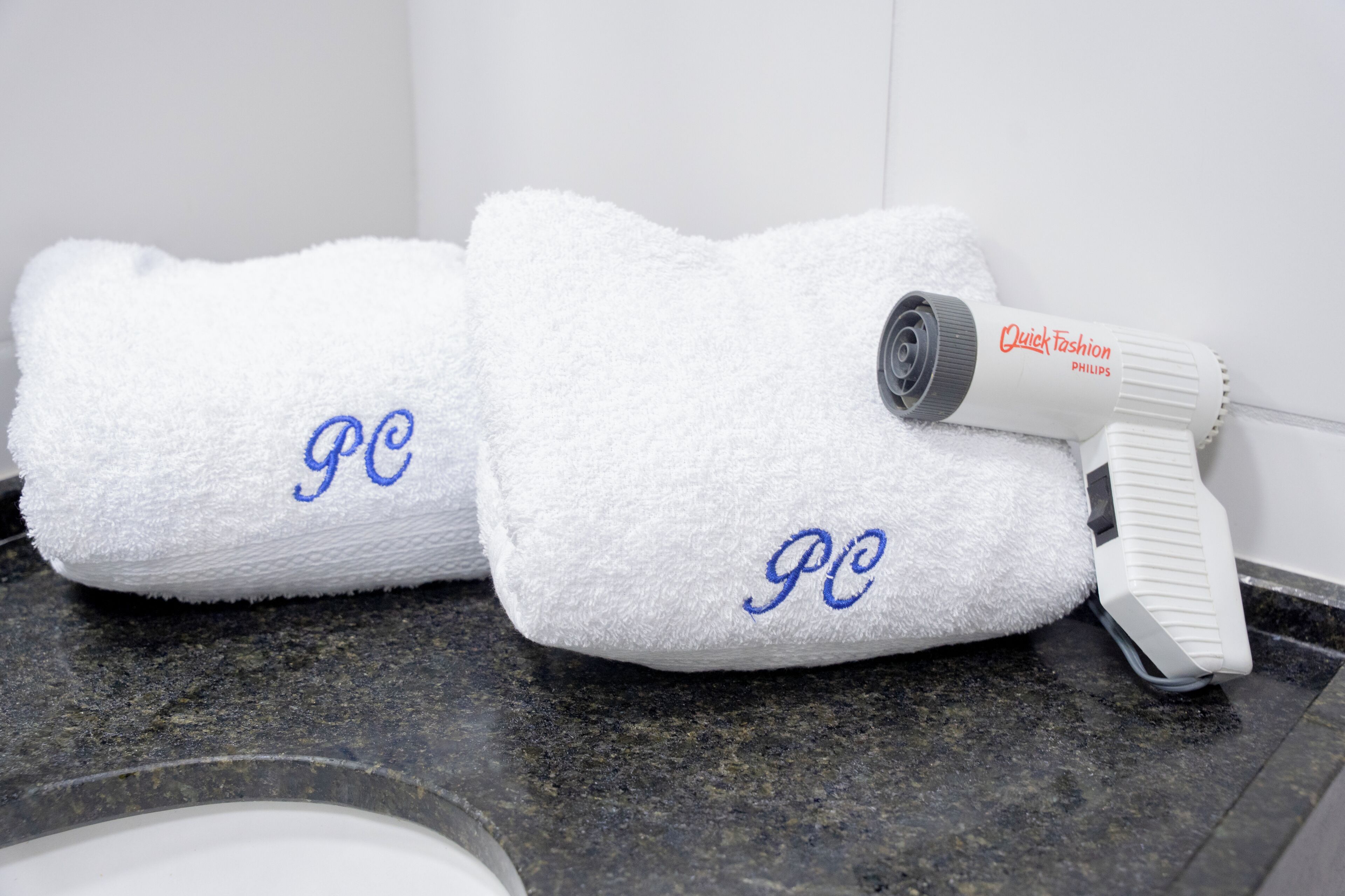 Two white towels with embroidered initials on a granite countertop, a hair dryer, and a white wall in the background.