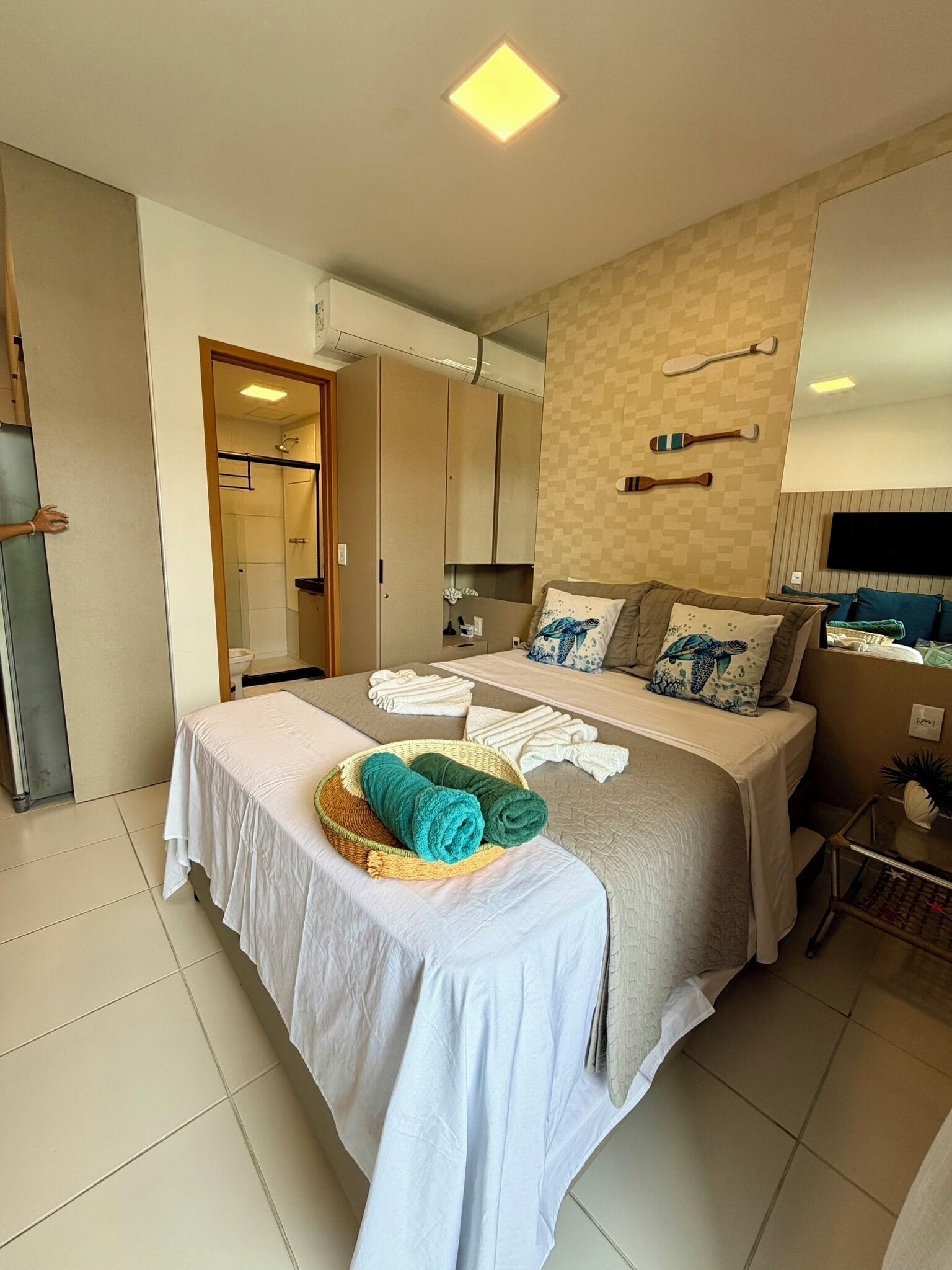 A modern hotel room with a bed, a bedside table, a wall-mounted TV, and a bathroom visible through an open door.
