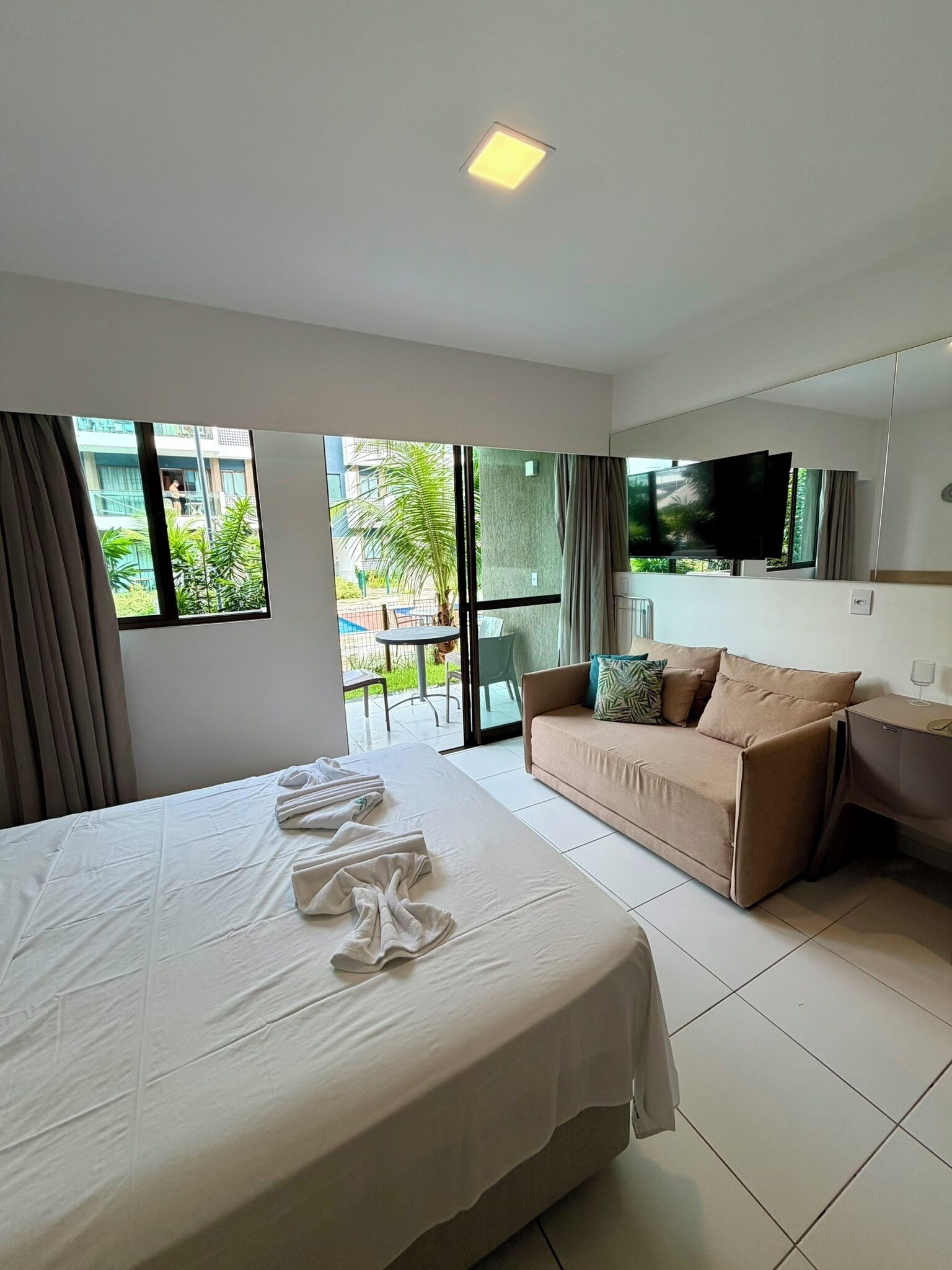 A hotel room with a large bed, a sofa, a TV, and a balcony with a view of greenery.