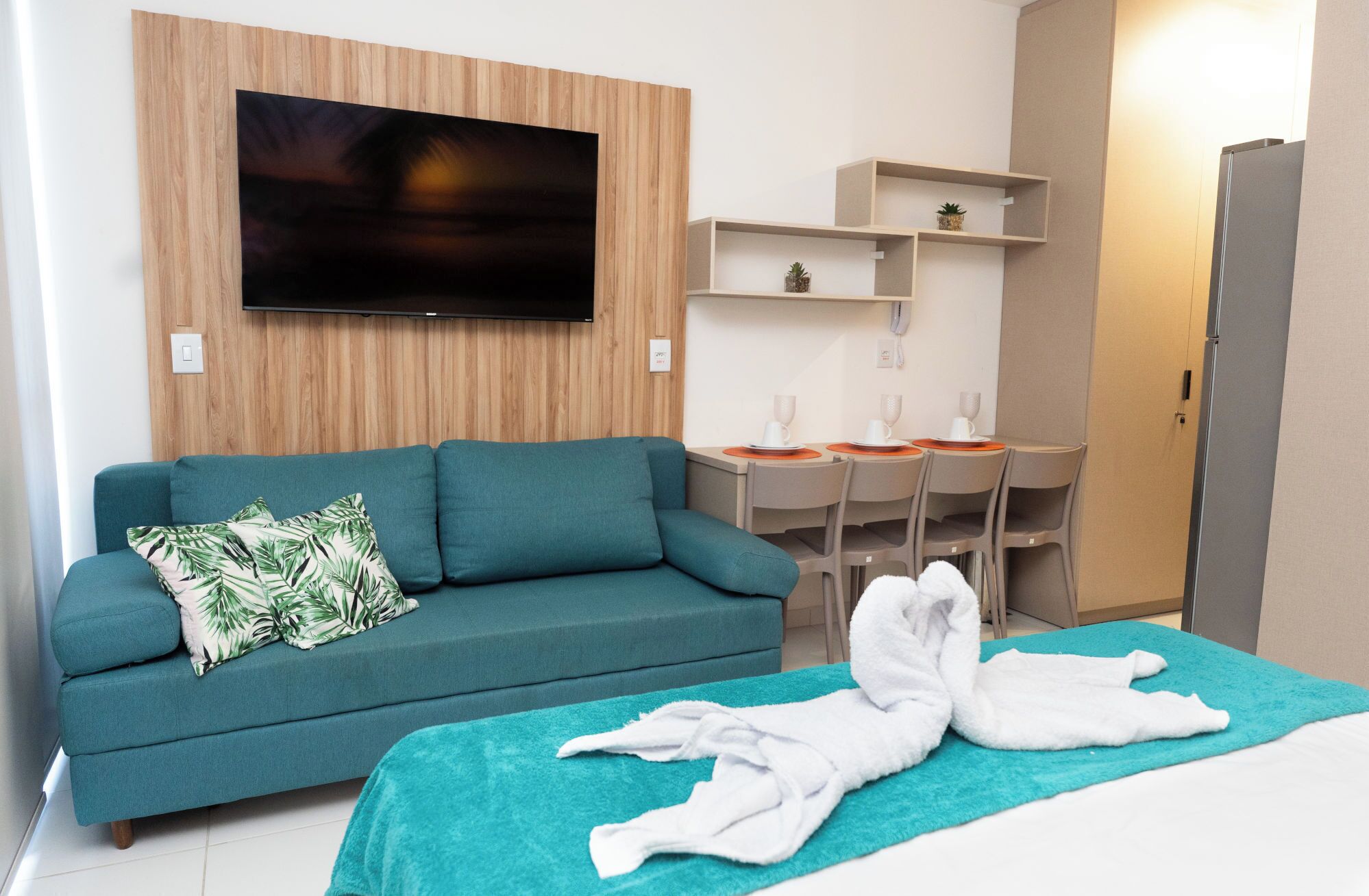 A hotel room with a teal sofa, a TV mounted on a wooden panel wall, a desk with chairs, and a bed with a teal blanket.