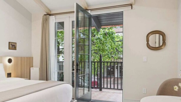 A hotel room with a bed, a chair, a round table, and a balcony with a view of greenery.