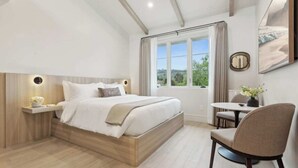 A modern bedroom with a large bed, bedside tables, a chair, a small table, and a window with curtains. - Casa Riviera 20 - Boutique Suites (Santa Barbara)