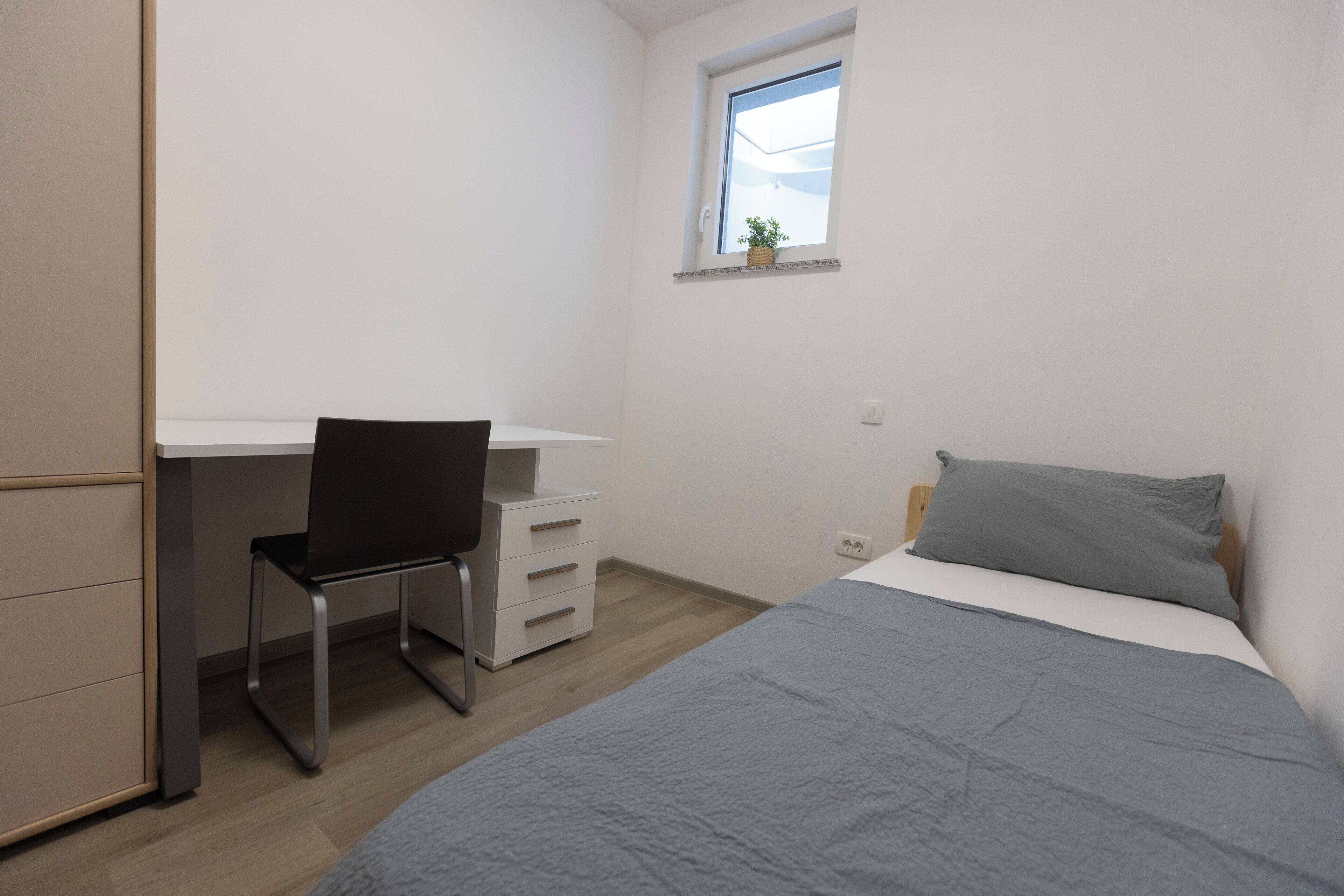 Un dormitorio con cama, escritorio, silla, cómoda y ventana con planta.