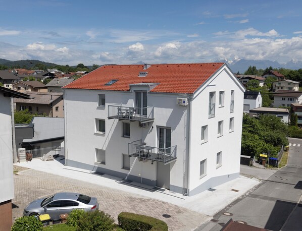A modern two-story building with a red-tiled roof, multiple balconies, and a parking area with a car and a yellow suitcase. - Villa Andrea Apartments - Ljubljana (Ljubljana)