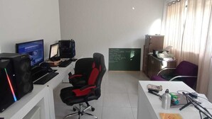 A small office with two desks, computers, a black office chair, a chalkboard, and a window with curtains. - ST DE PARNAIBA APARTMENT (Chácara do Solar II Santana de Parnaíba)