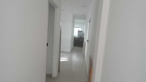 A narrow, well-lit hallway with white walls and a tiled floor, leading to a kitchen area. - ST DE PARNAIBA APARTMENT (Chácara do Solar II Santana de Parnaíba)