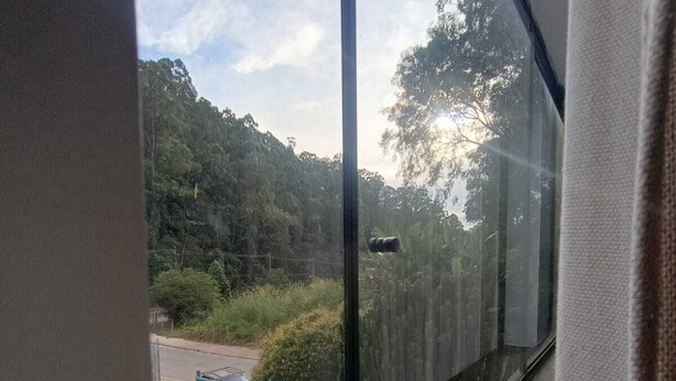 A room with a large window offering a view of a forested area and a road. - ST DE PARNAIBA APARTMENT (Chácara do Solar II Santana de Parnaíba)