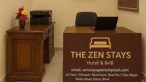 A reception desk with a laptop, a printer, and a vase of flowers. There is a television mounted on the wall displaying a corridor. A wooden statue is placed above the desk, and a signboard for "The Zen Stays Hotel & BRI" is visible. - The Zen Stays (Danapur)