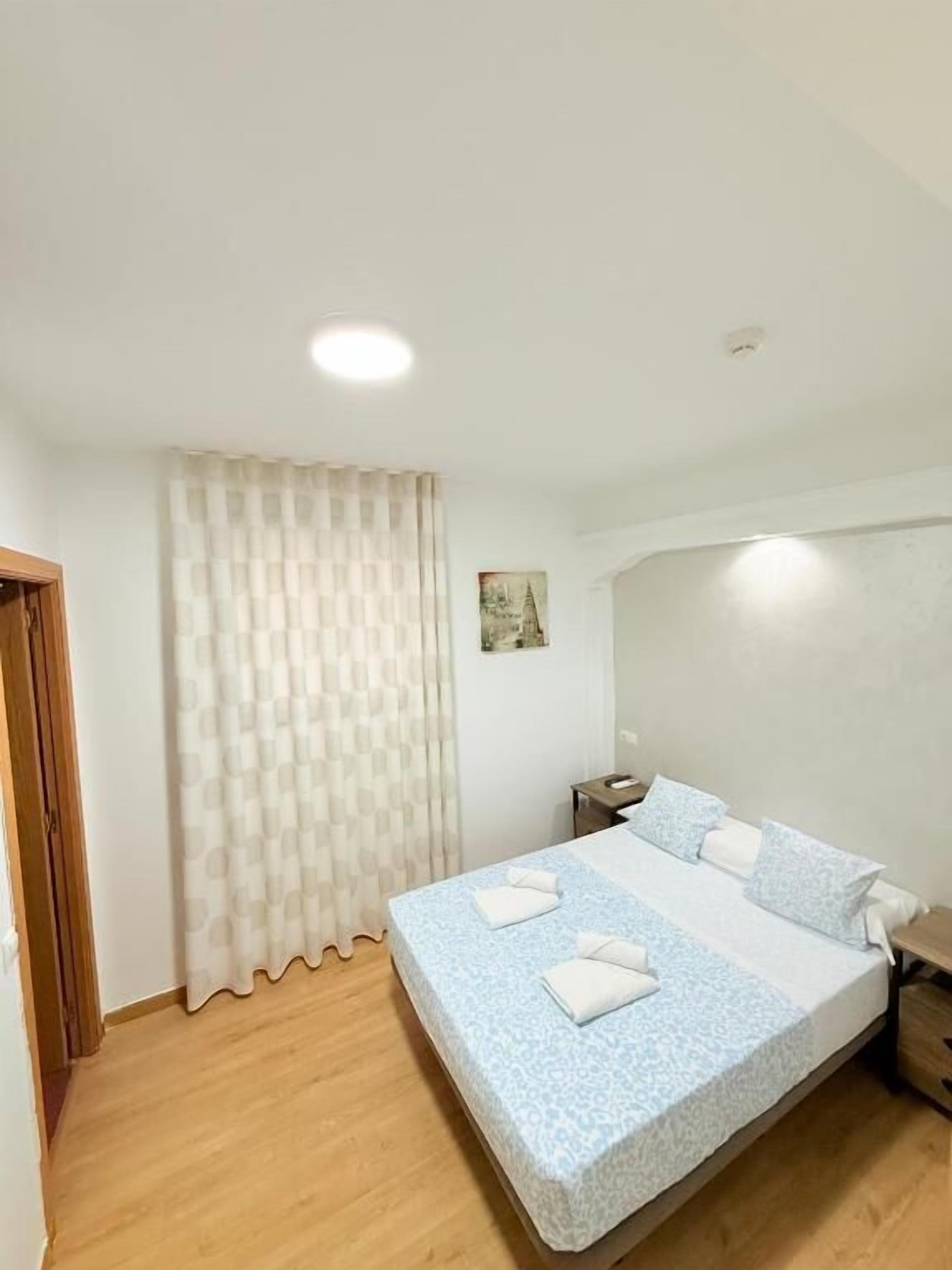 A bedroom with a bed, a nightstand, a wall-mounted light, a window with curtains, and a door.
