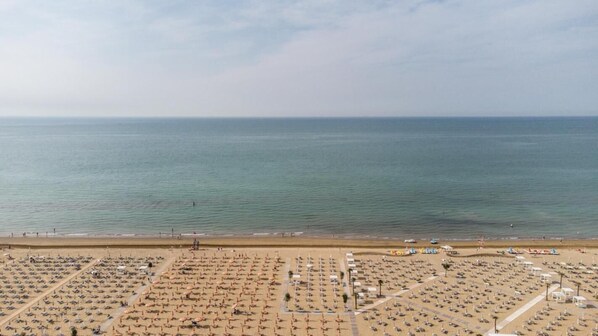 Eine Vogelperspektive zeigt einen Strand mit zahlreichen Liegestühlen und Sonnenschirmen, einem wolkenlosen Himmel und ruhiger See.