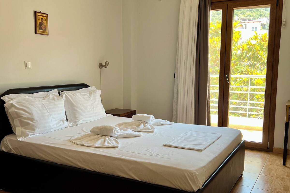 A neatly made bed with white linens and towels, a wooden headboard, a small bedside table, and a wall-mounted air conditioner.