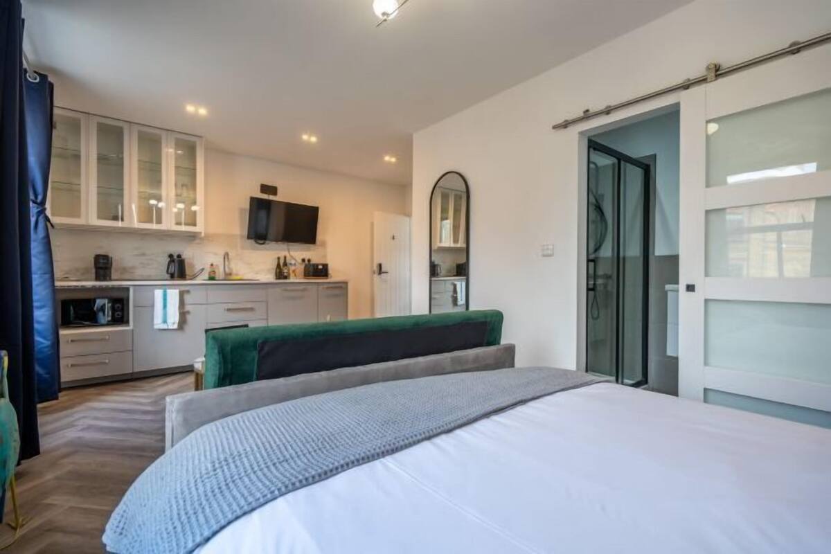 A modern bedroom with a large bed, a green bench, a TV, a kitchen area, and a glass door leading to a bathroom.
