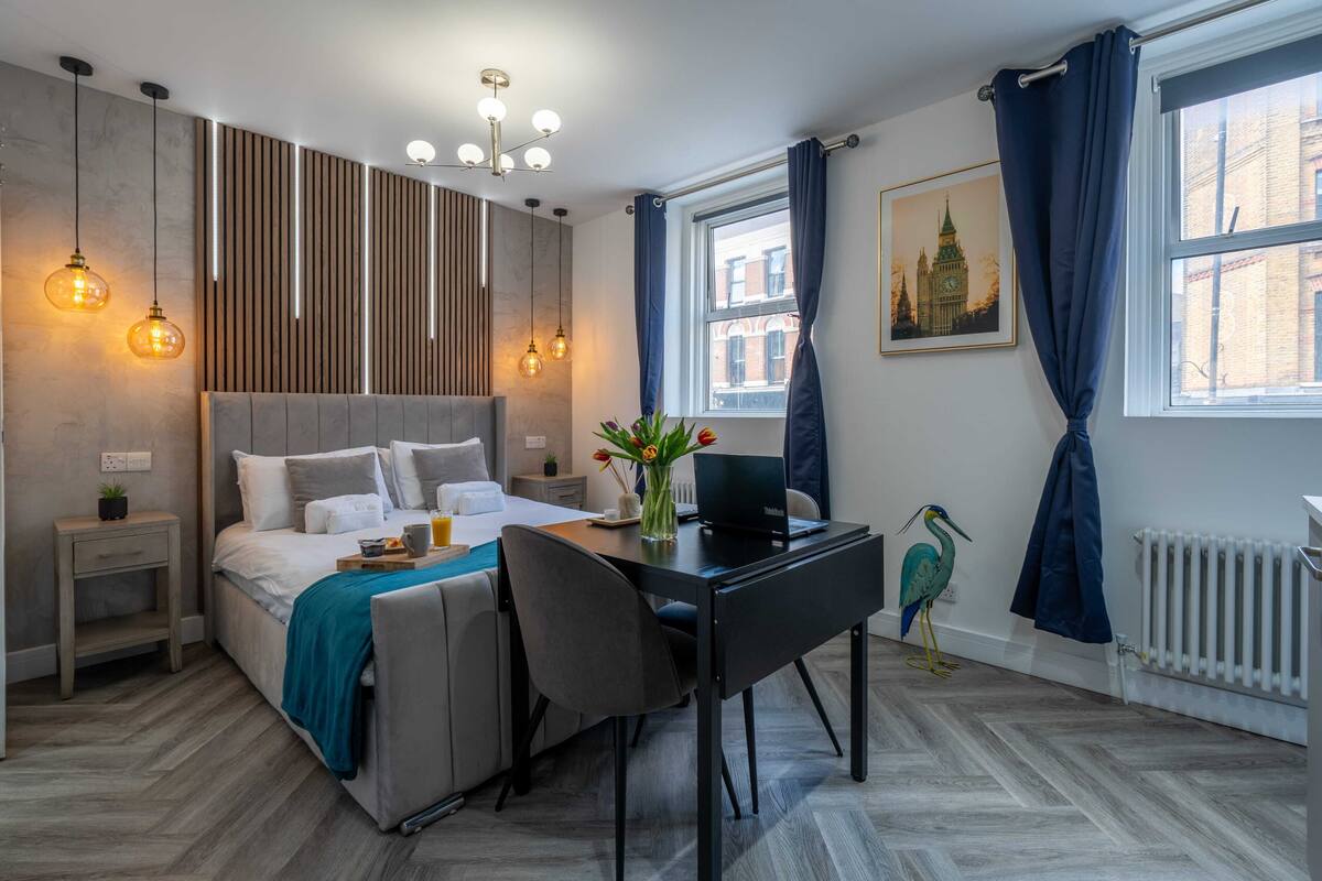 A modern hotel room with a bed, desk, and chair, a window with curtains, a radiator, and a wall-mounted light fixture.