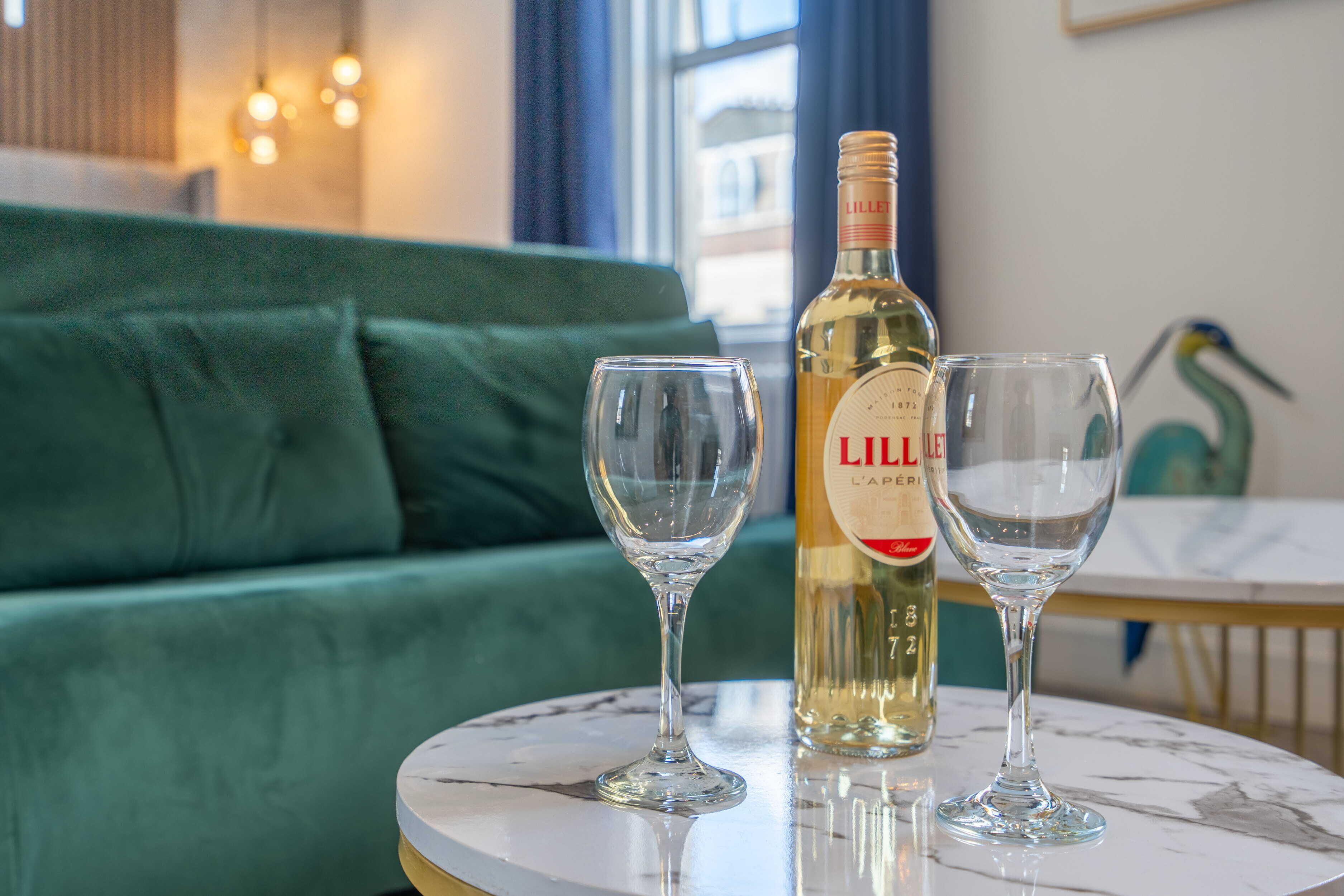 Una botella de Lillet y dos copas sobre una mesa de mármol en una habitación con un sofá verde y una ventana con cortinas.