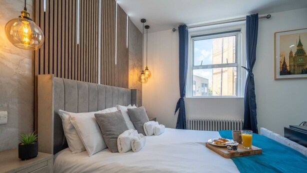 A neatly made bed with pillows and a tray of breakfast, a window with curtains, and a wall-mounted light fixture. - Shoreditch Luxe (London)