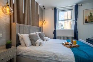 A neatly made bed with pillows and a tray of breakfast, a window with curtains, and a wall-mounted light fixture. - Shoreditch Luxe (London)