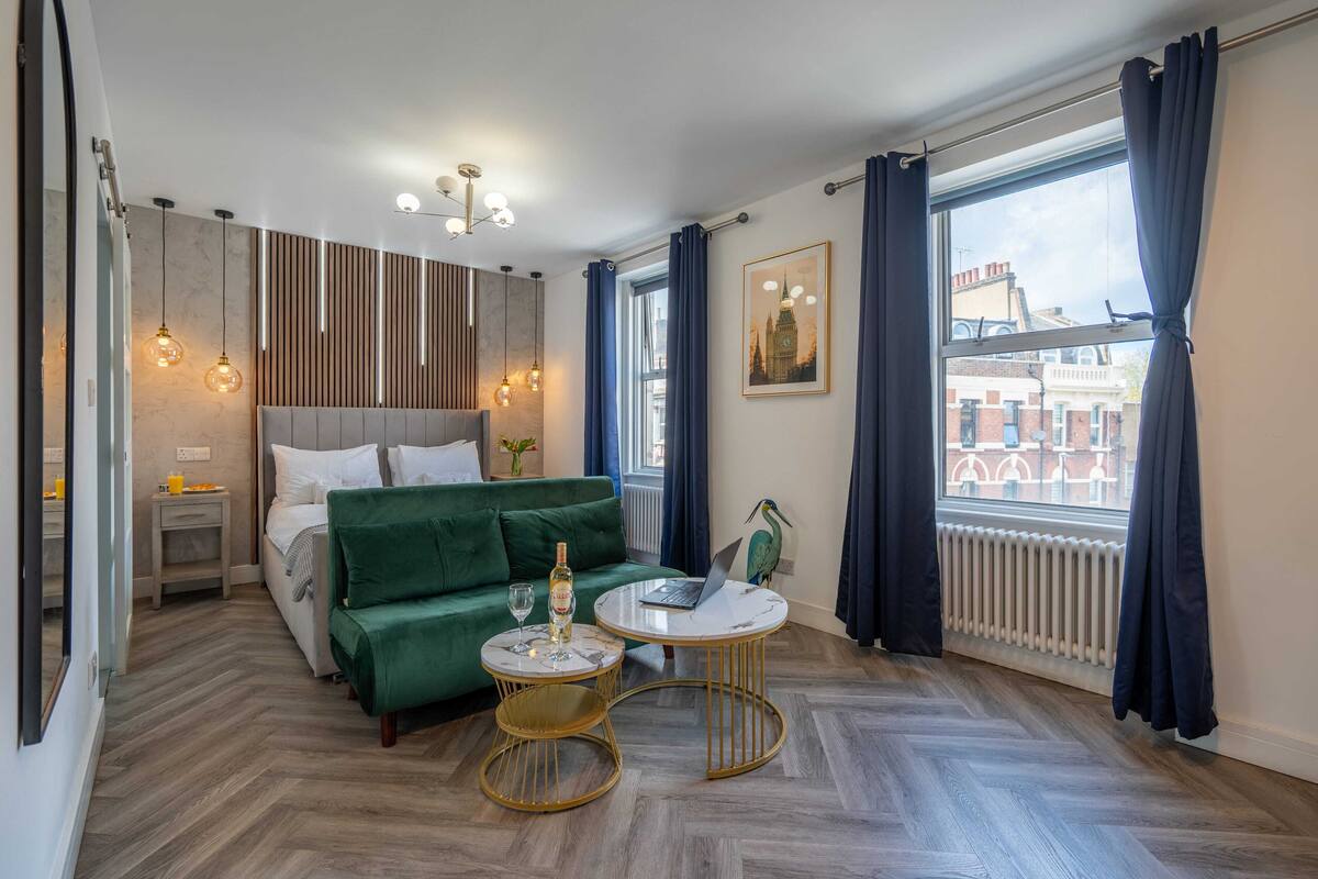 A modern bedroom with a green sofa, a round table with a laptop, and a view of buildings through the window.