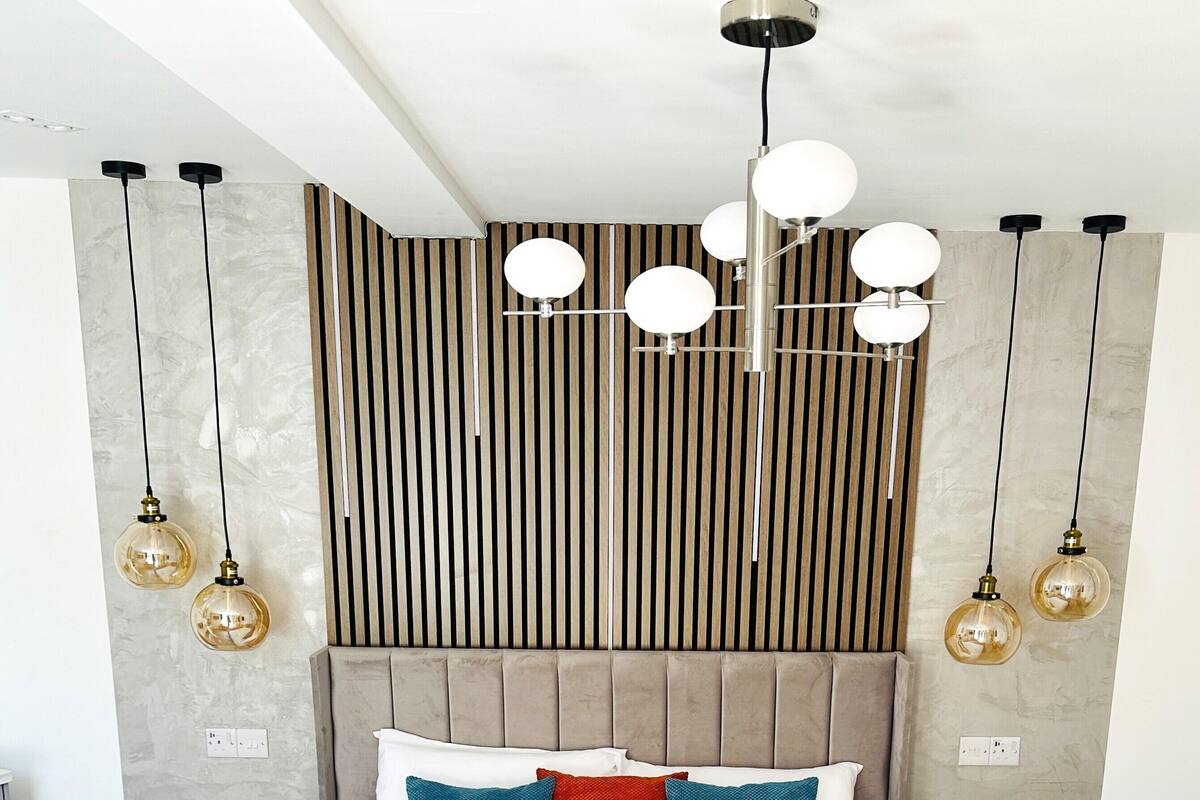A modern bedroom with a tufted headboard, a large bed with white and red bedding, two bedside tables, and hanging pendant lights.