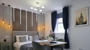 A modern bedroom with a bed, bedside tables, a desk with chairs, a window with curtains, and a wall-mounted light fixture.