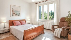 A bedroom with a wooden bed, a chair, a window with a view of greenery, a radiator, and a painting on the wall. - Beautiful home in Sablet (Sablet)