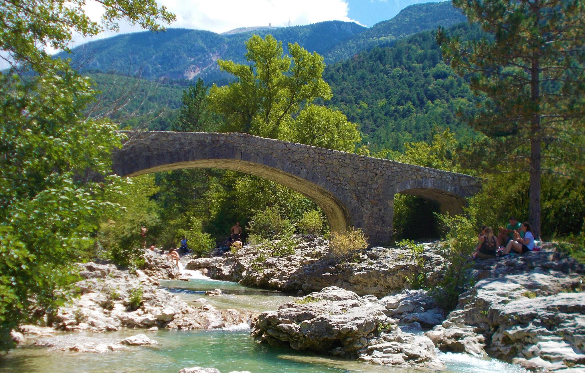 Un puente de piedra sobre un río, con gente sentada sobre las rocas, rodeado de vegetación y montañas.