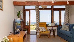 A living room with a TV on a wooden stand, a blue sofa, a yellow armchair, and a view of the sea through large windows. - Historic Railway Home - Selsey Seafront (Chichester)
