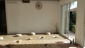Een ruime kamer met een hoge plafond, grote ramen en een houten vloer. Er liggen vloerkleden en kussens voor zitplekken of om op te bewegen.