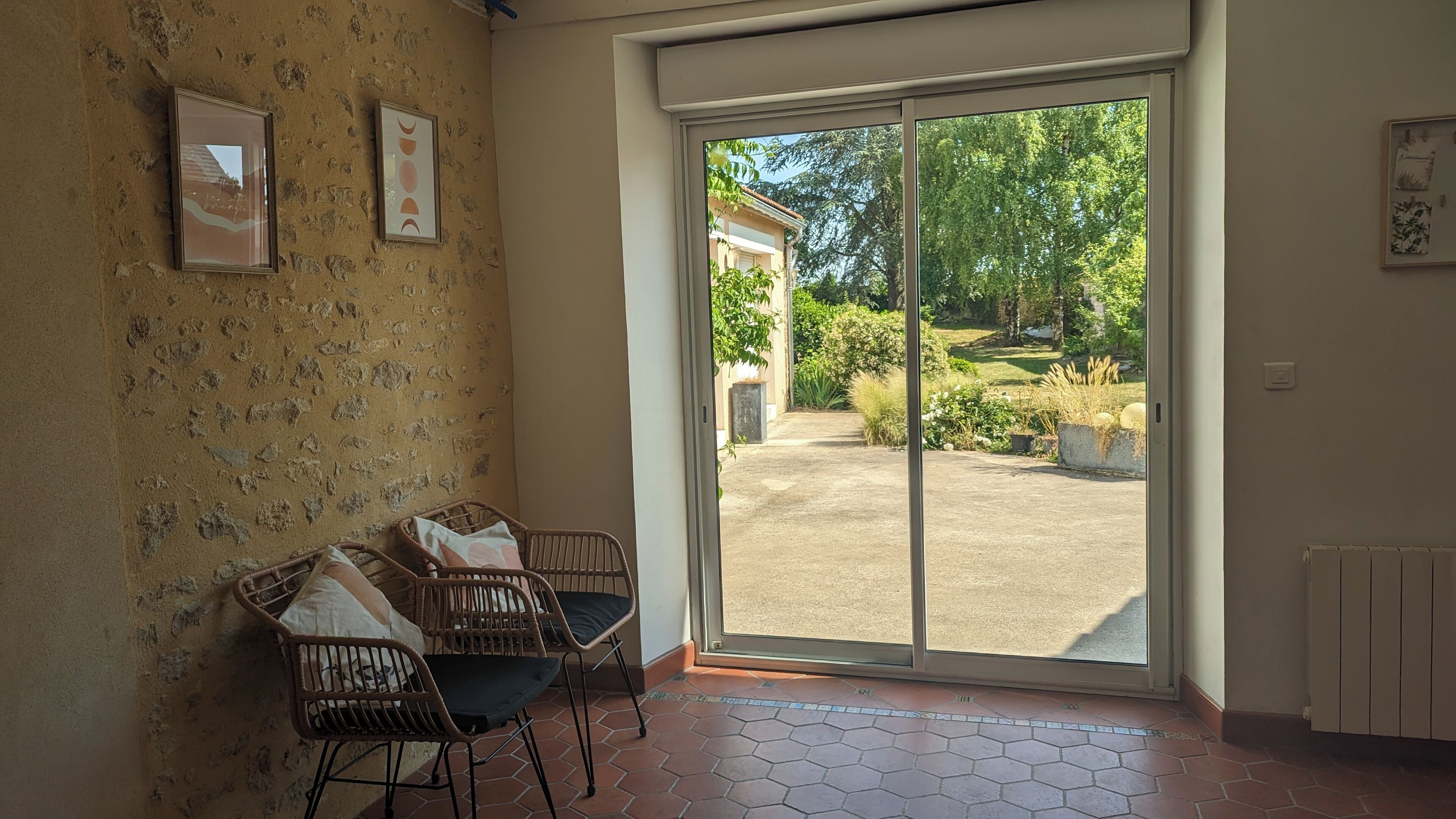 A room with a large sliding glass door leading to a garden, wicker chairs, and a tiled floor.