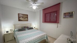 A bedroom with a bed, nightstands, a ceiling fan, a window with blinds, a wall-mounted picture, and a small table with a decorative item.