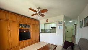 A living room with wooden cabinets, a television, a ceiling fan, a kitchen area with a microwave, and a sofa.