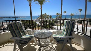 A balcony with two striped chairs and a glass table, overlooking a beach with palm trees and the ocean.