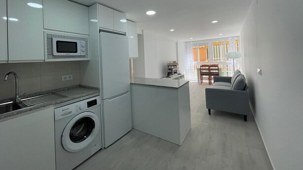 A modern kitchen with a built-in washing machine, a microwave, and a refrigerator. There is a dining area with a table and chairs visible through a window. - TERRASOL MONTERREY 2ªB7 (Torre del Mar)