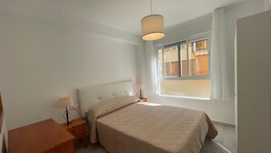 A bedroom with a large bed, a bedside table, a lamp, and a window with a view of buildings. - TERRASOL MONTERREY 2ªB7 (Torre del Mar)