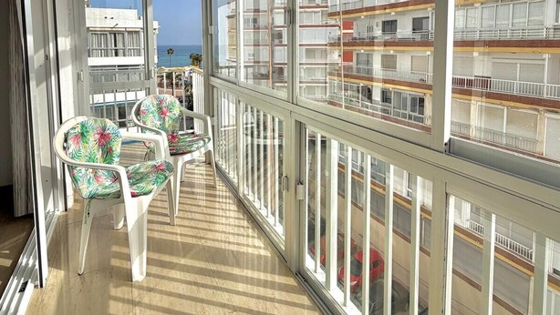 A balcony with large windows, white chairs, and a view of buildings and the sea. - TERRASOL SAN FRANCISCO 5-2 (Torre del Mar)
