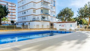 A swimming pool with a tiled deck, surrounded by a white building with balconies and a small structure with a sign. - TERRASOL SAN FRANCISCO 5-2 (Torre del Mar)
