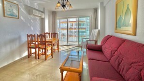 A living room with a glass-top coffee table, a red sofa, wooden chairs, a chandelier, and a balcony with a view of buildings. - TERRASOL SAN FRANCISCO 5-2 (Torre del Mar)