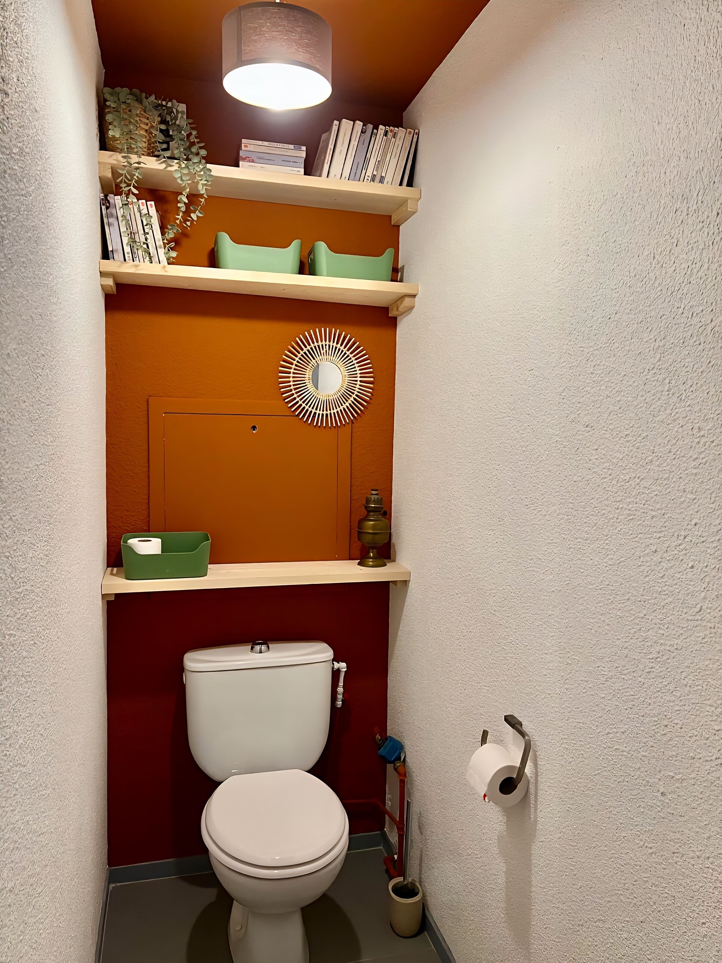 Een compacte badkamer met een wit toilet, planken met boeken en toiletartikelen, en een decoratief wandobject.