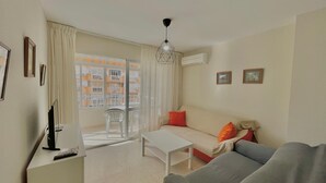 A living room with a sofa, a small table, a TV, and a balcony with a view of buildings. - TERRASOL CAPRI 2 (Torre del Mar)