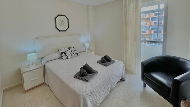 A bedroom with a bed, bedside tables, a black leather chair, a window with curtains, and a cityscape view. - TERRASOL CAPRI 2 (Torre del Mar)
